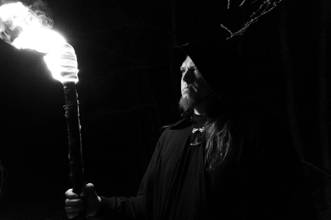 T. M. Mäkelä in a hooded cloak holding a burning torch in a dark forest. Photo by TikSu